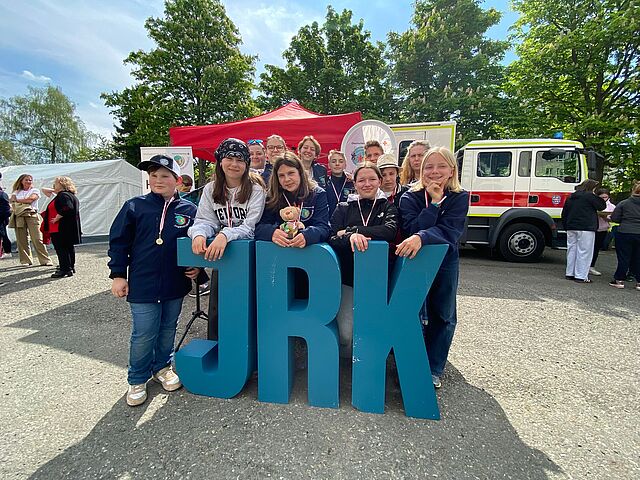 Eine Gruppe junger Menschen steht draußen hinter großen blauen JRK-Buchstaben; im Hintergrund sind ein rotes Zelt und ein Fahrzeug zu sehen.