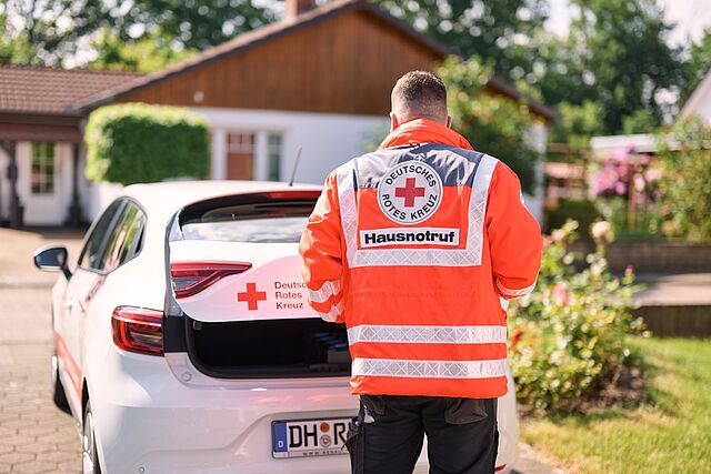 Person in orangefarbener DRK-Warnjacke hinter einem weißen Auto mit geöffneter Heckklappe