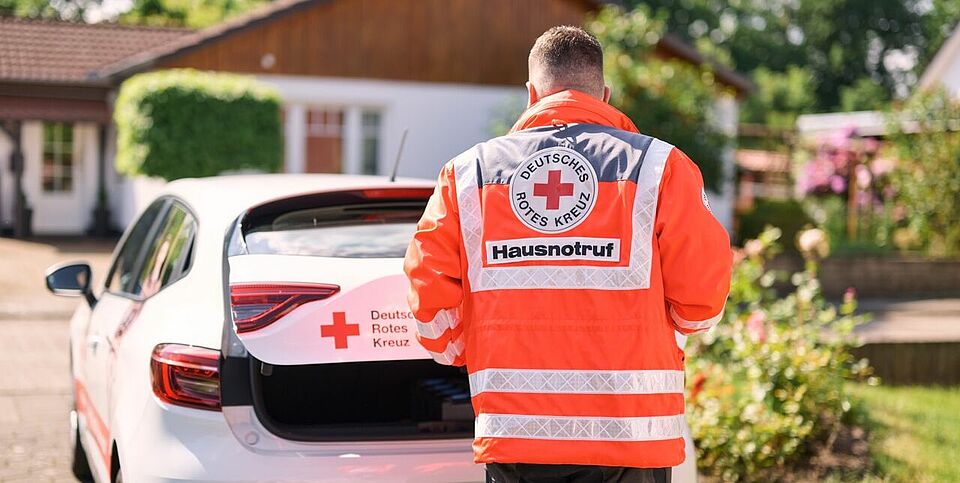 Person in orangefarbener DRK-Warnjacke hinter einem weißen Auto mit geöffneter Heckklappe