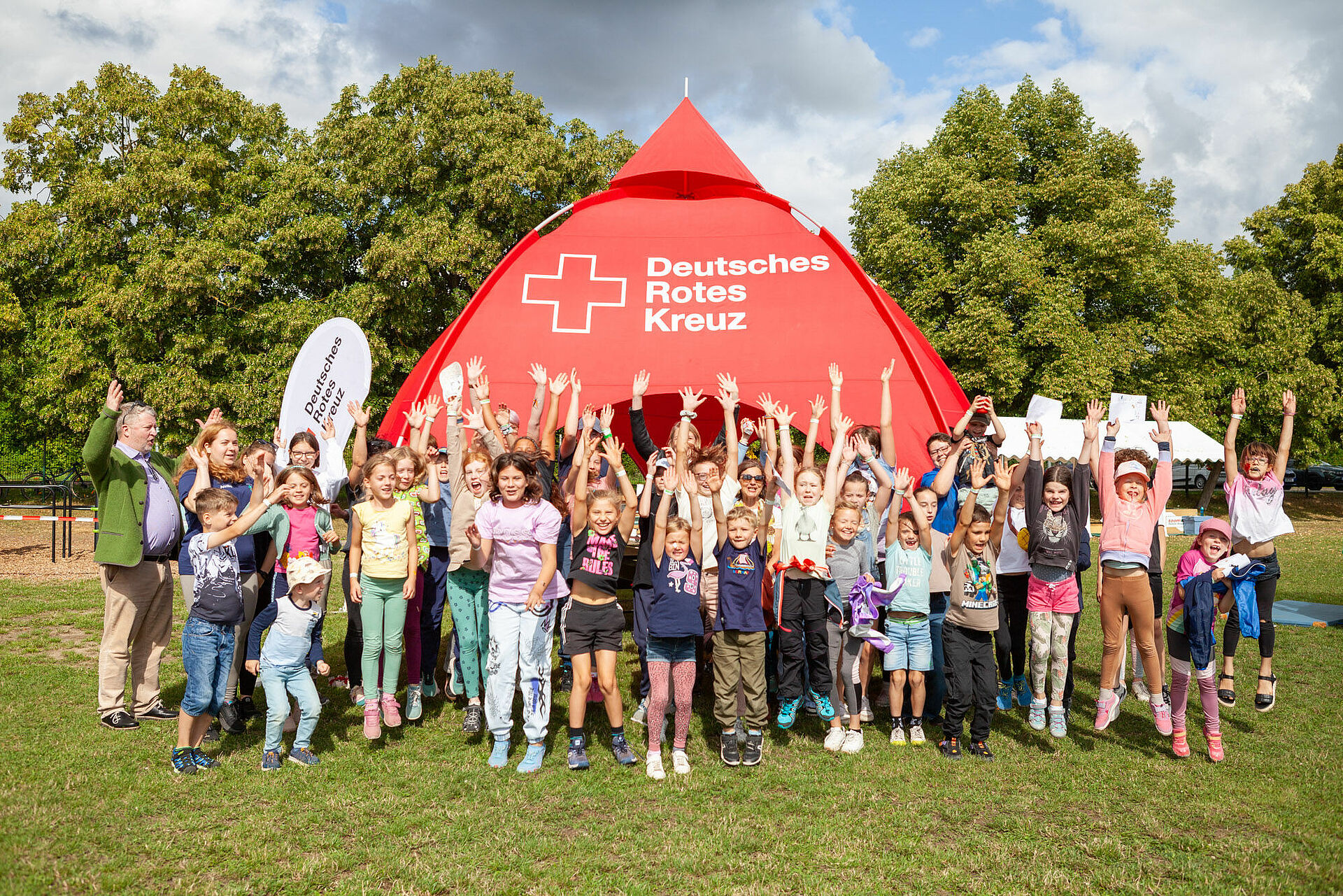 Große Kindergruppe jubelt mit erhobenen Armen vor einem roten Zelt mit Logo des Deutschen Roten Kreuzes auf einer Wiese