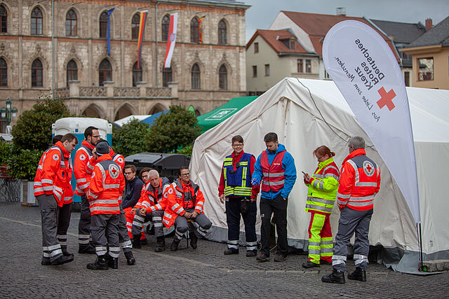 Mehrere Menschen in roten und gelben Warnjacken stehen auf einer Straße vor einem Gebäude.