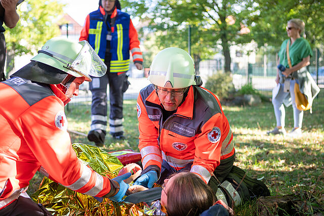 Zwei Einsatzkräfte des Deutschen Roten Kreuzes in orangefarbener Schutzkleidung und Helmen versorgen eine auf dem Boden liegende Person unter einer goldenen Rettungsdecke.